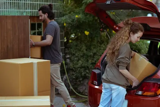 Focused young woman with curly hair taking cardboard package out from red automobile trunk on street near stack of moving boxes while bearded ethnic man carrying box into new house