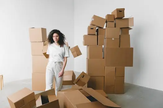 A young woman stands indoors surrounded by stacked cardboard boxes.